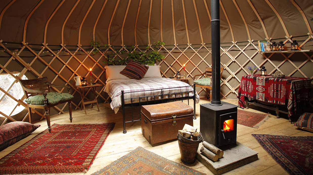 A cozy, rustic bedroom styled inside a yurt. Yellow walls, metal-framed bed, fire in the stove. Kunderi kilim rugs are spr...